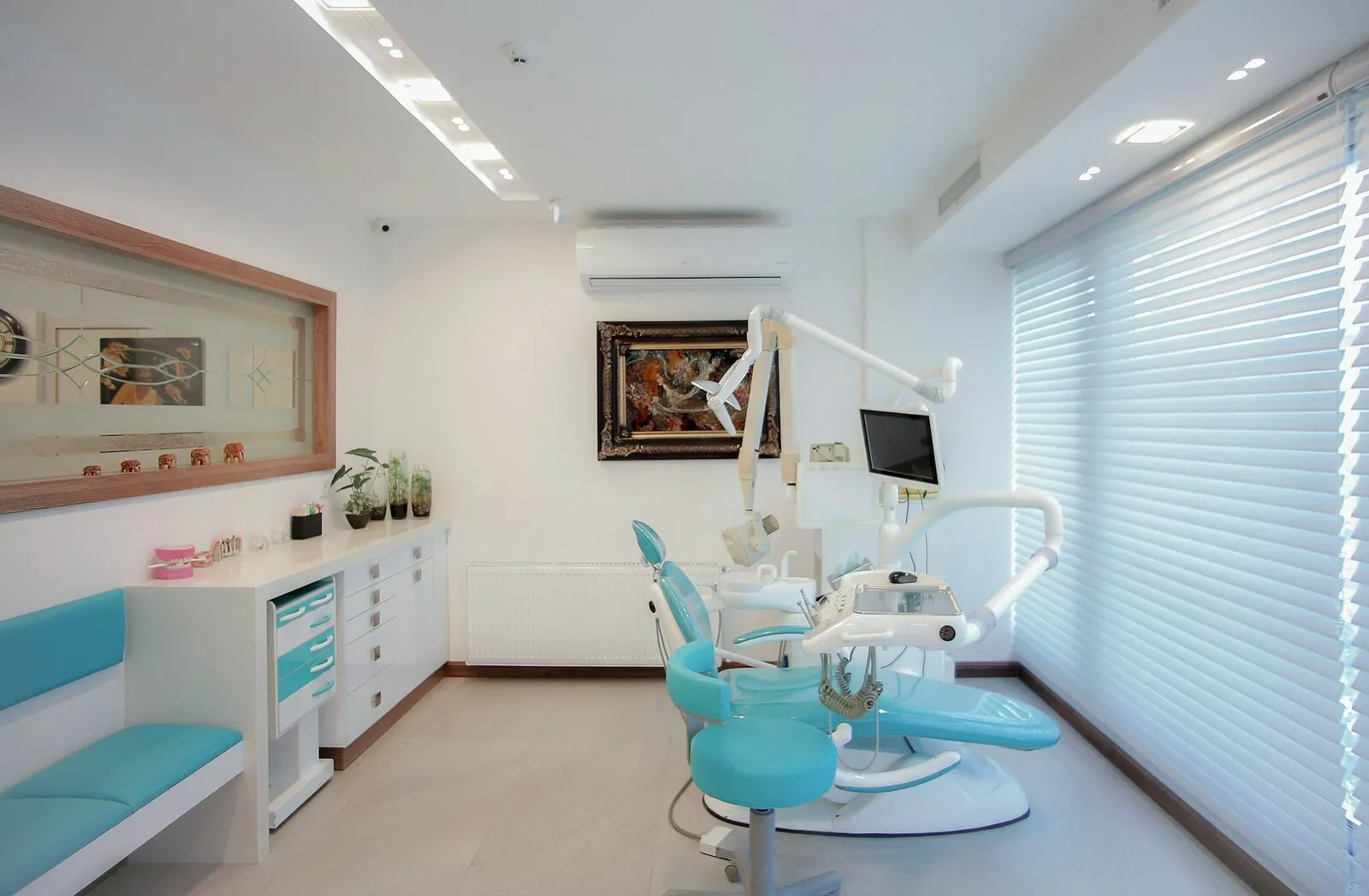 A bright, empty dental treatment room with a teal chair and a window with blinds